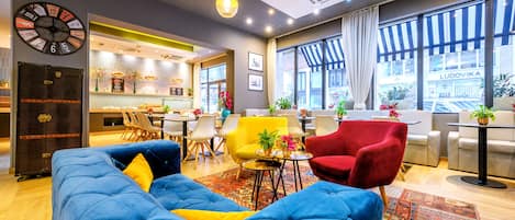 A modern hotel lobby with a blue sofa, red chairs, a yellow armchair, and a large clock on the wall.