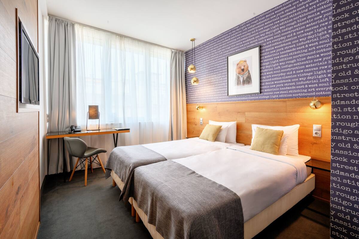 A hotel room with two beds, a desk, a chair, and a large window with curtains.