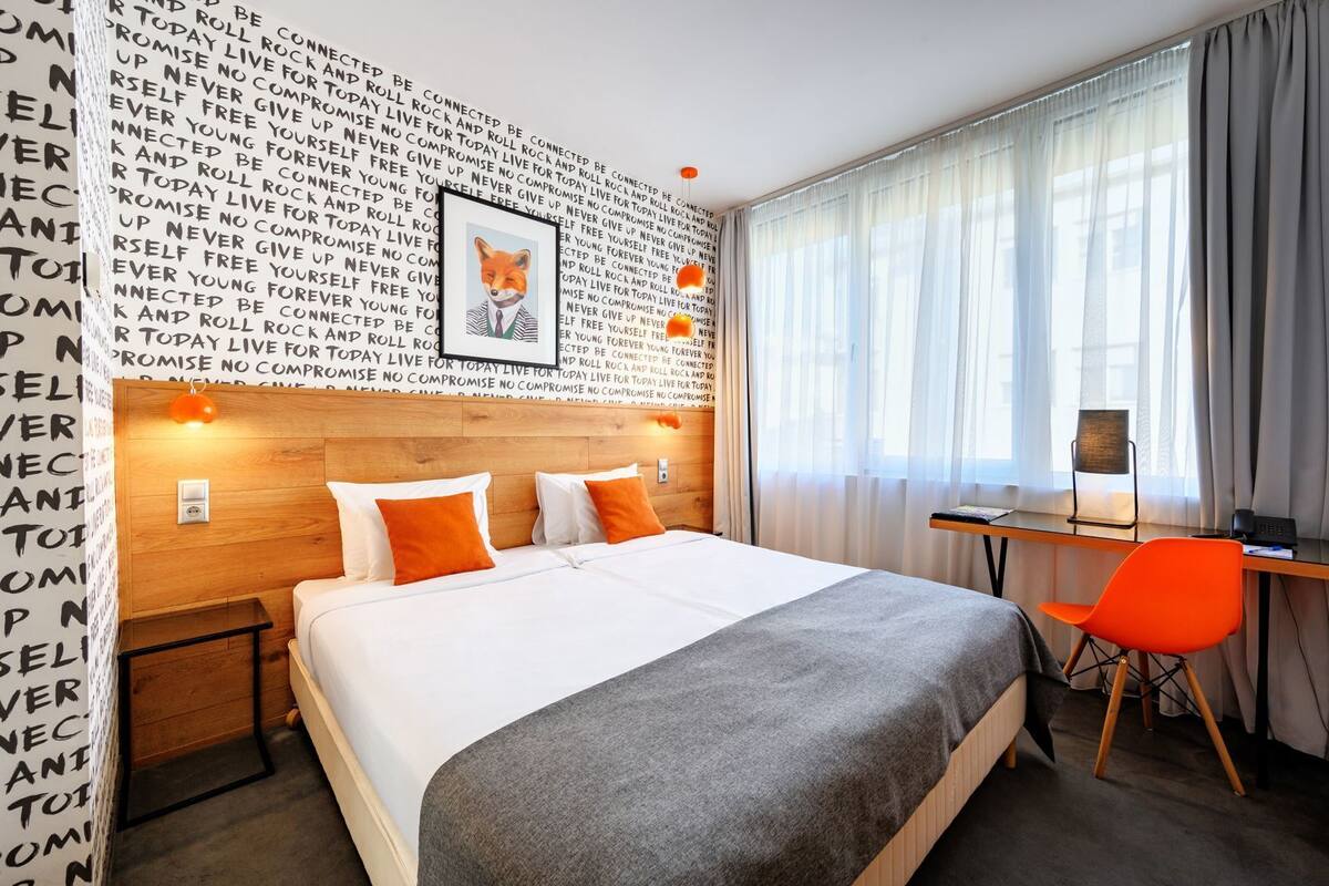 A hotel room with a large bed, a desk with a chair, a window with curtains, and a wall with repeating text.