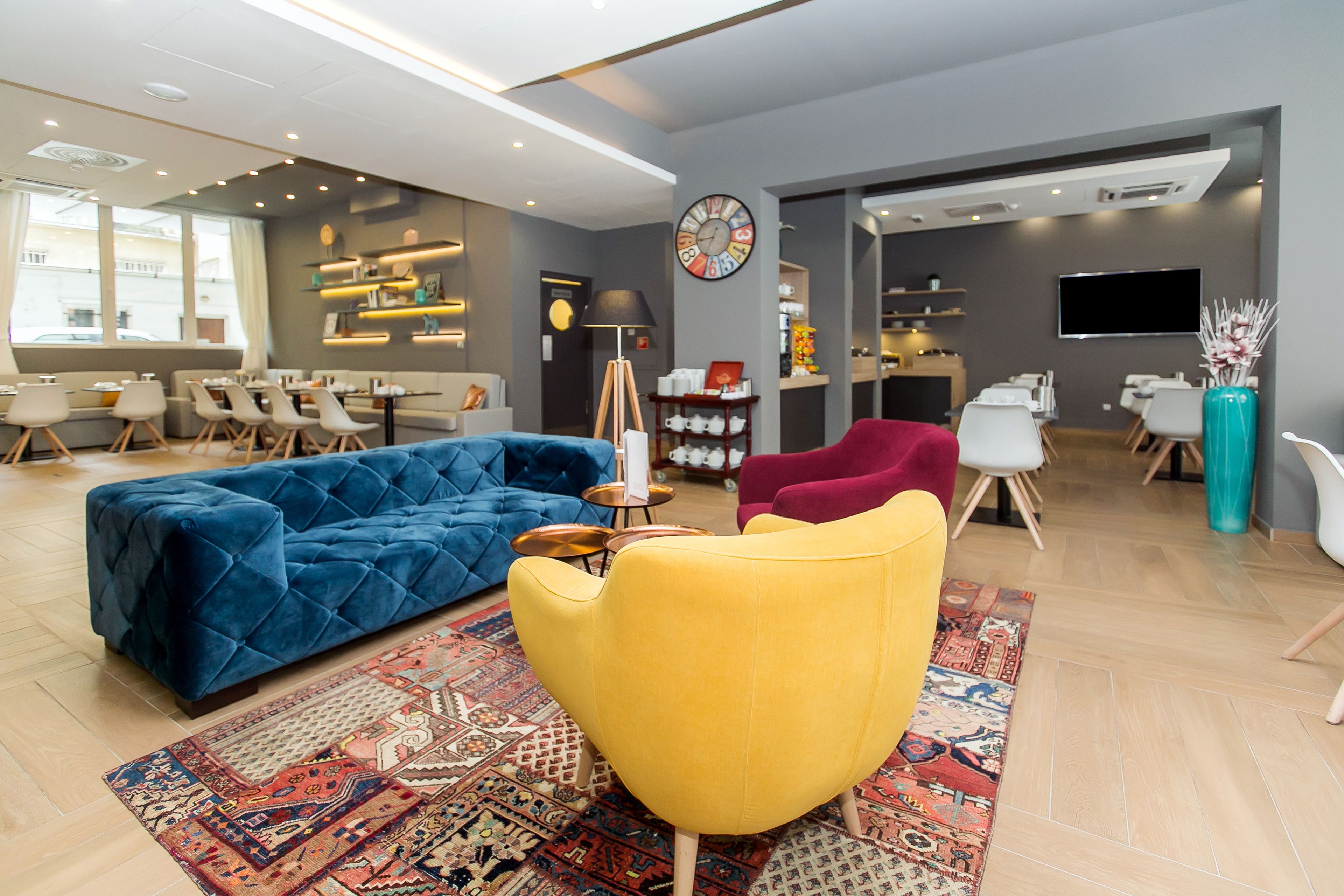 Un moderno vestíbulo de hotel con un sofá azul, un sillón amarillo y una silla roja. Hay mesas y sillas para comer, un reloj y un televisor en la pared.