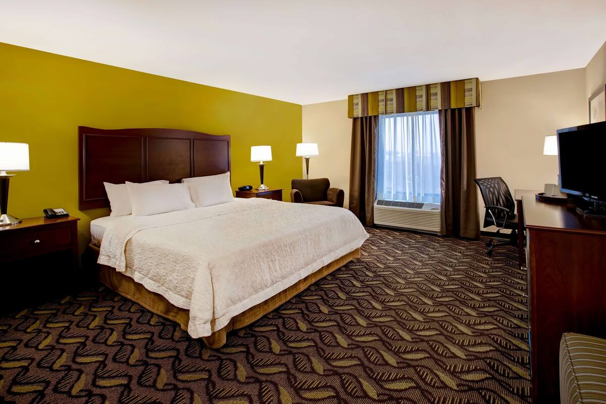 A hotel room with a large bed, two bedside tables, a desk with a chair, a television, and a window with curtains.