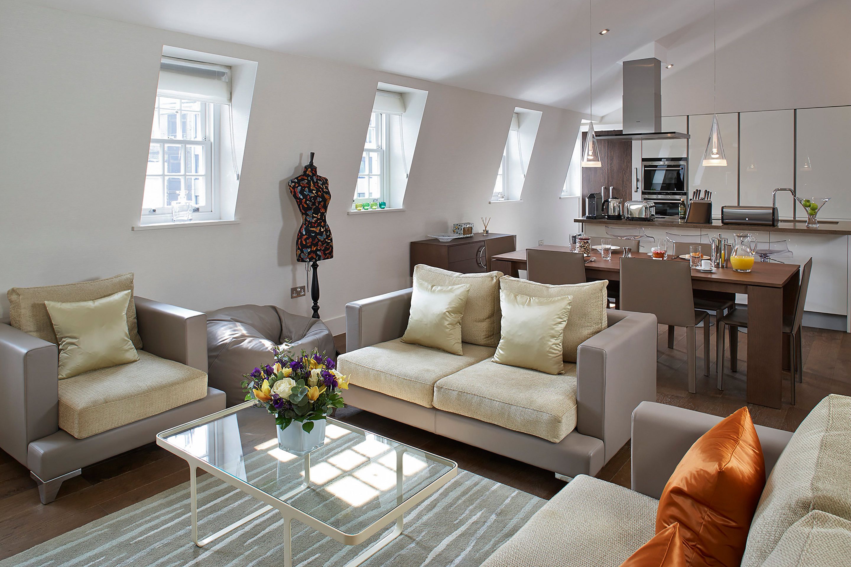A modern living room with a sofa, armchairs, and a glass coffee table. There is a kitchen area with a dining table and chairs.