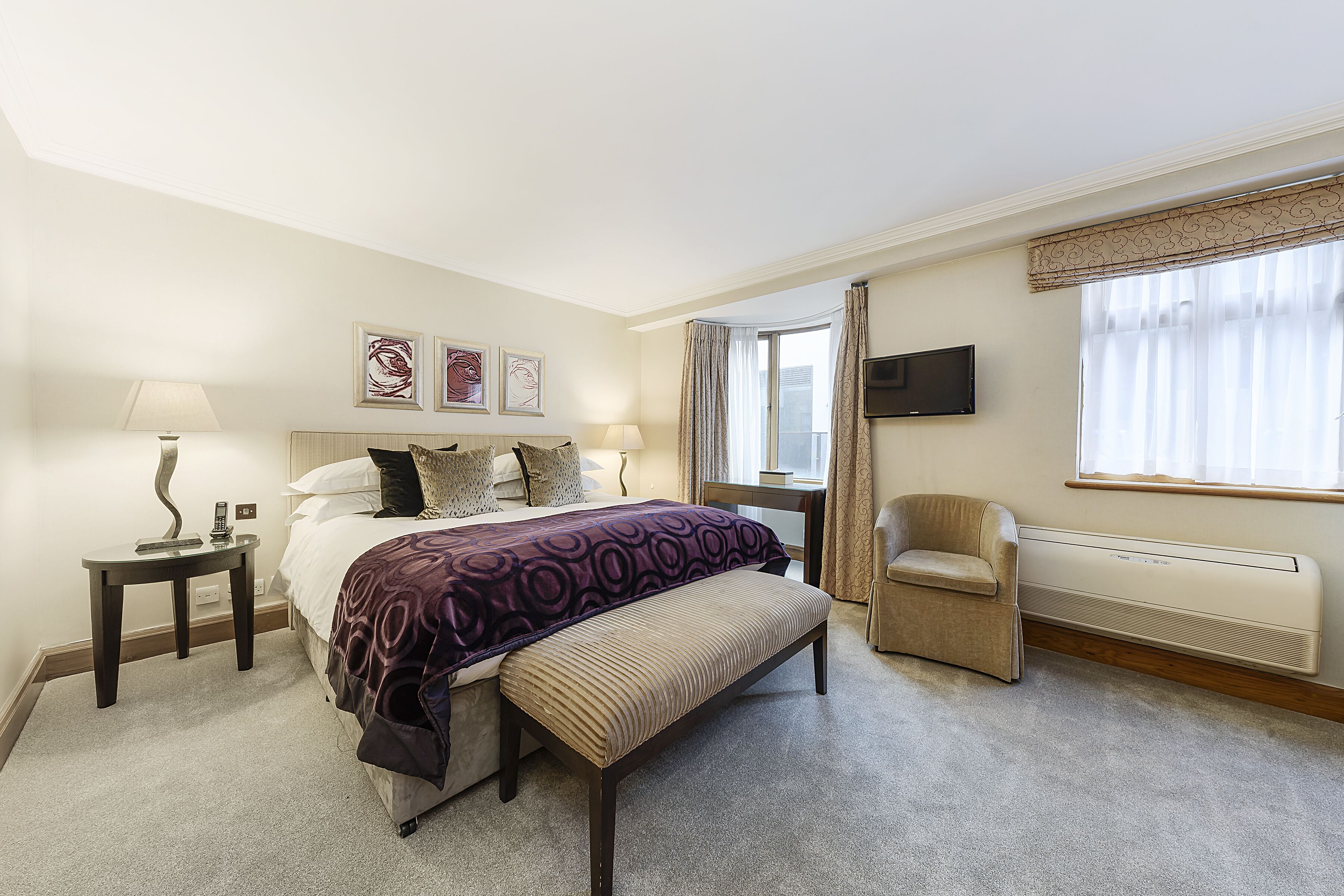 A hotel room with a bed, a bench, a chair, a desk, a TV, and a window with curtains.