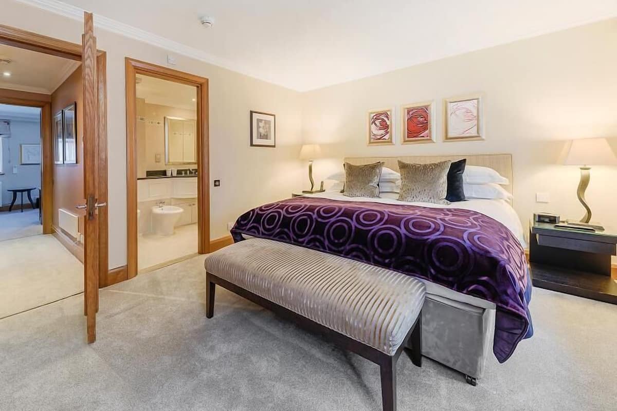 A bedroom with a bed, a bench, a nightstand, and a bathroom visible through an open door.