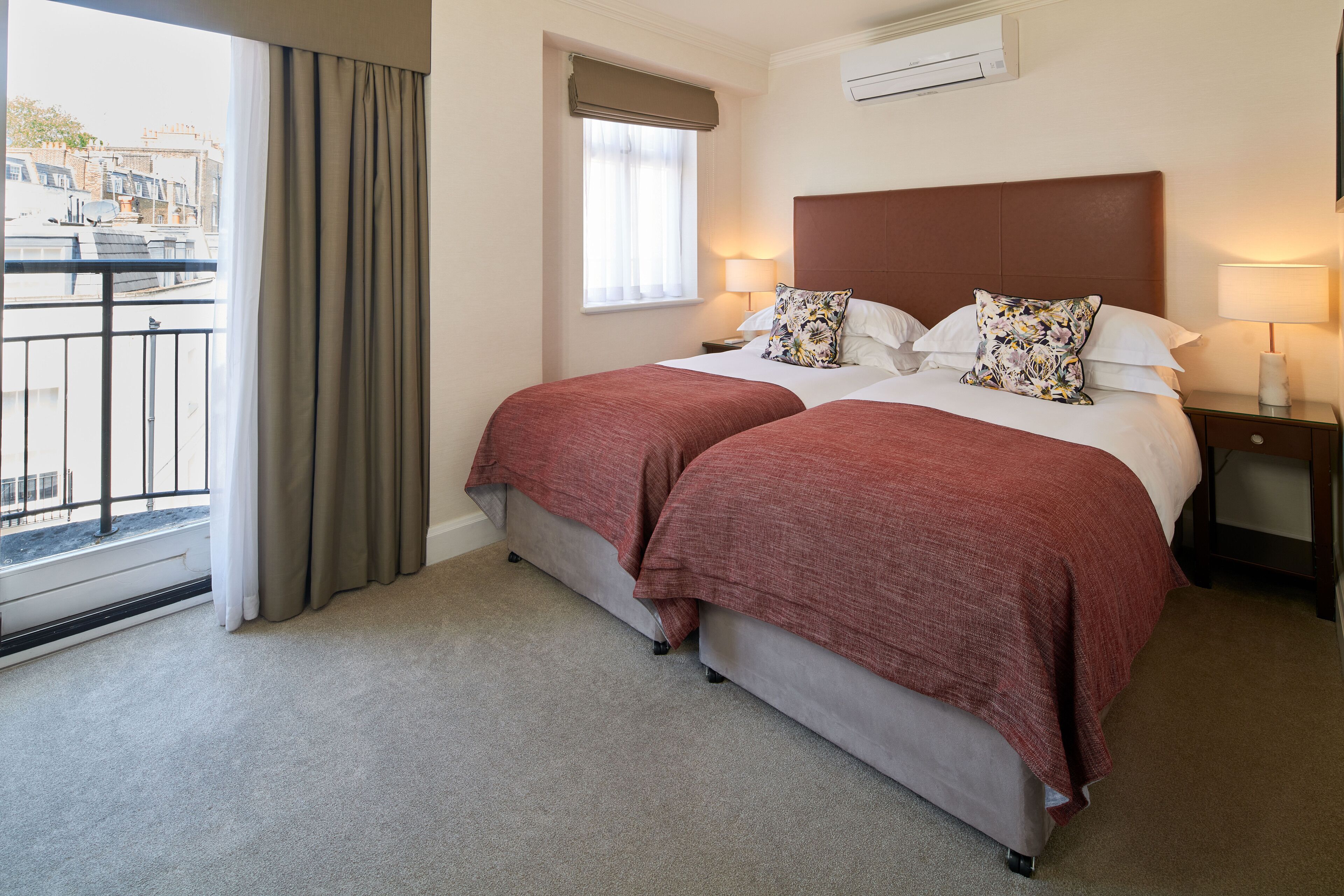 Een tweepersoonsbed met twee kussens, een nachtkastje met een lamp, een raam met gordijnen en een balkon met een hekwerk.
