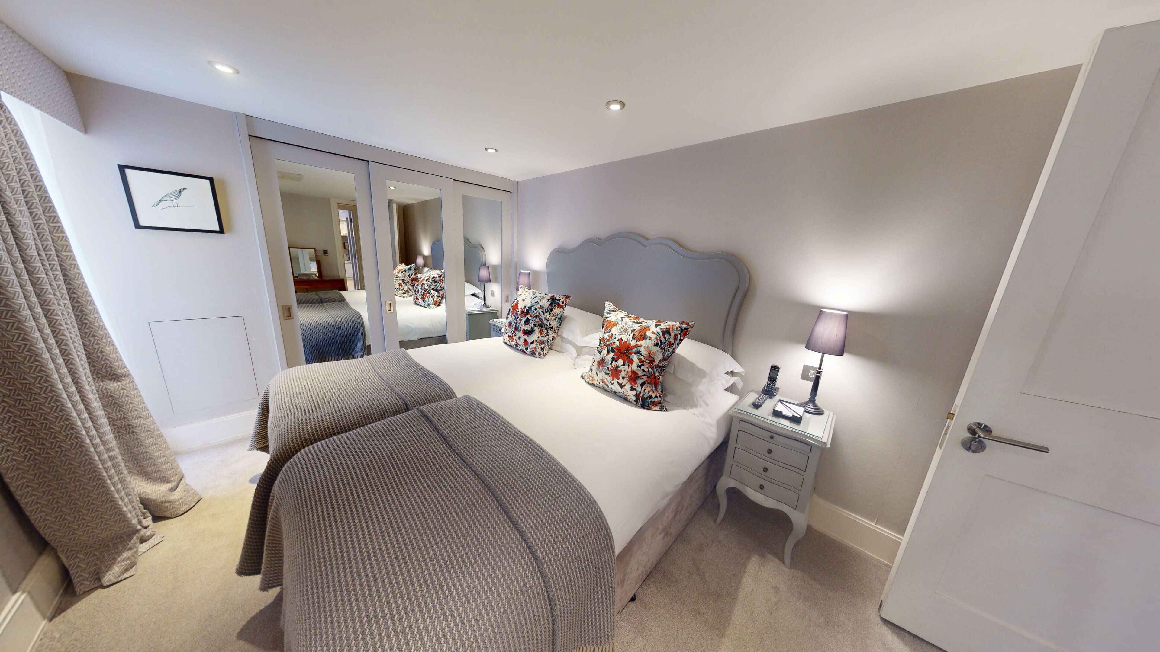 A bedroom with a large bed, two pillows, a bedside table with a lamp, a mirror, and a wardrobe.