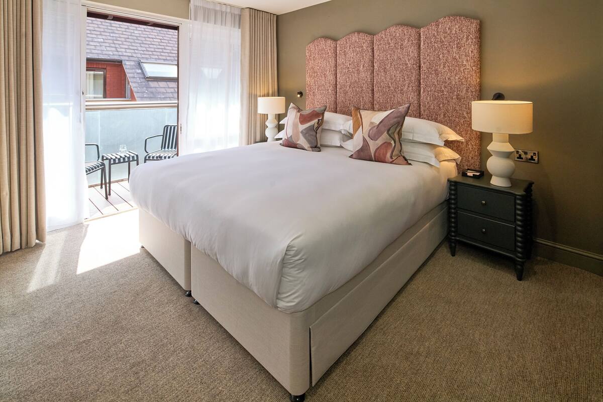 A hotel room with a large bed, two bedside tables with lamps, a view of the outside balcony, and a patterned headboard.