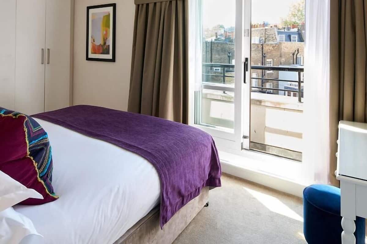 A hotel room with a bed, a window with curtains, a door, and a view of buildings outside.