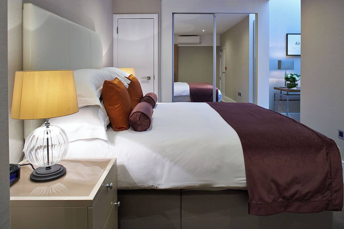A hotel room with a bed, a bedside lamp, a mirror, and a small table with a lamp.
