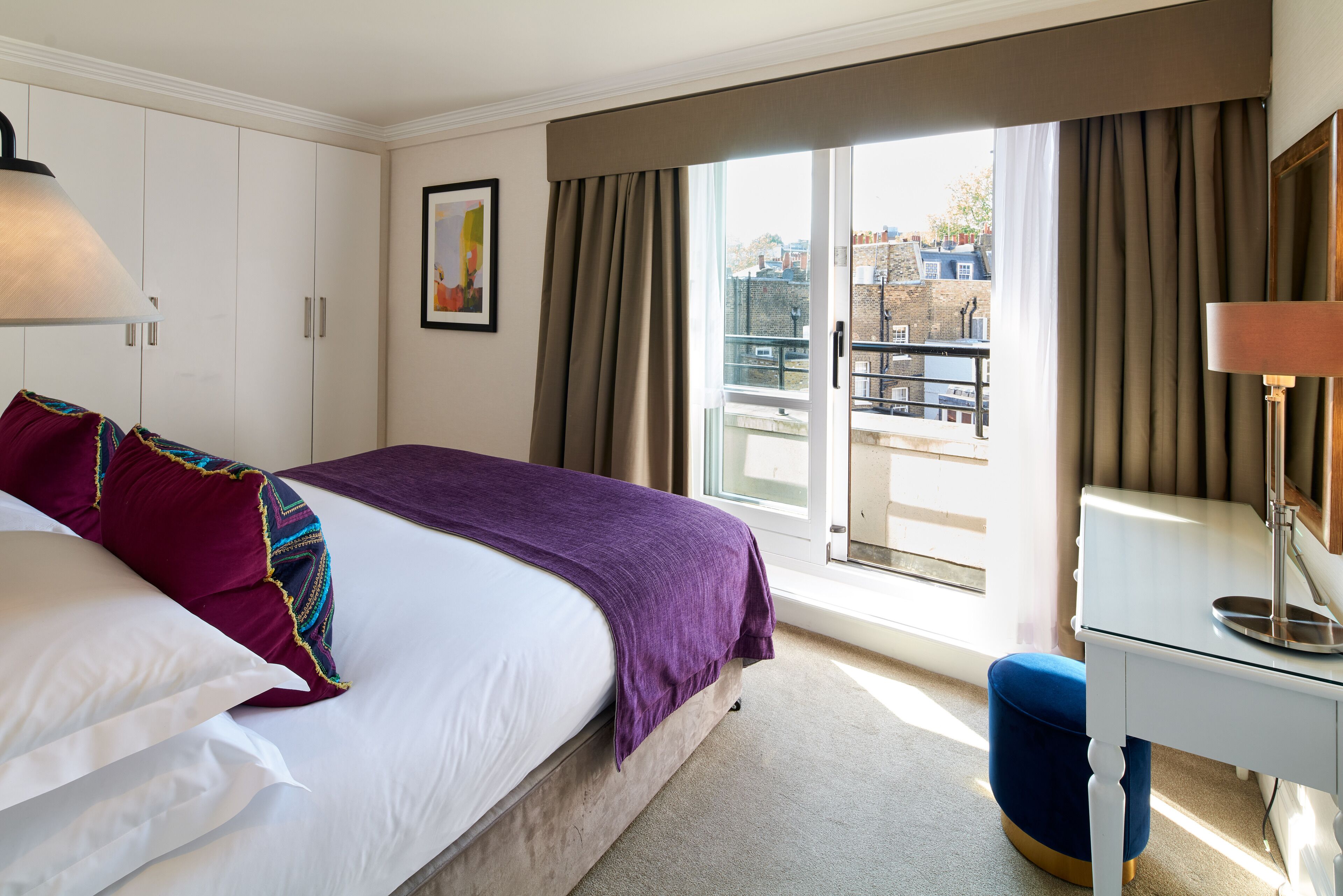 Habitación de hotel con cama, escritorio, silla, ventana con cortinas y vista a la ciudad.