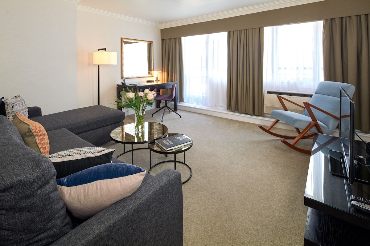 A modern hotel room with a grey sofa, a small round coffee table with a vase, a desk with a chair, a TV, and a large window with curtains.