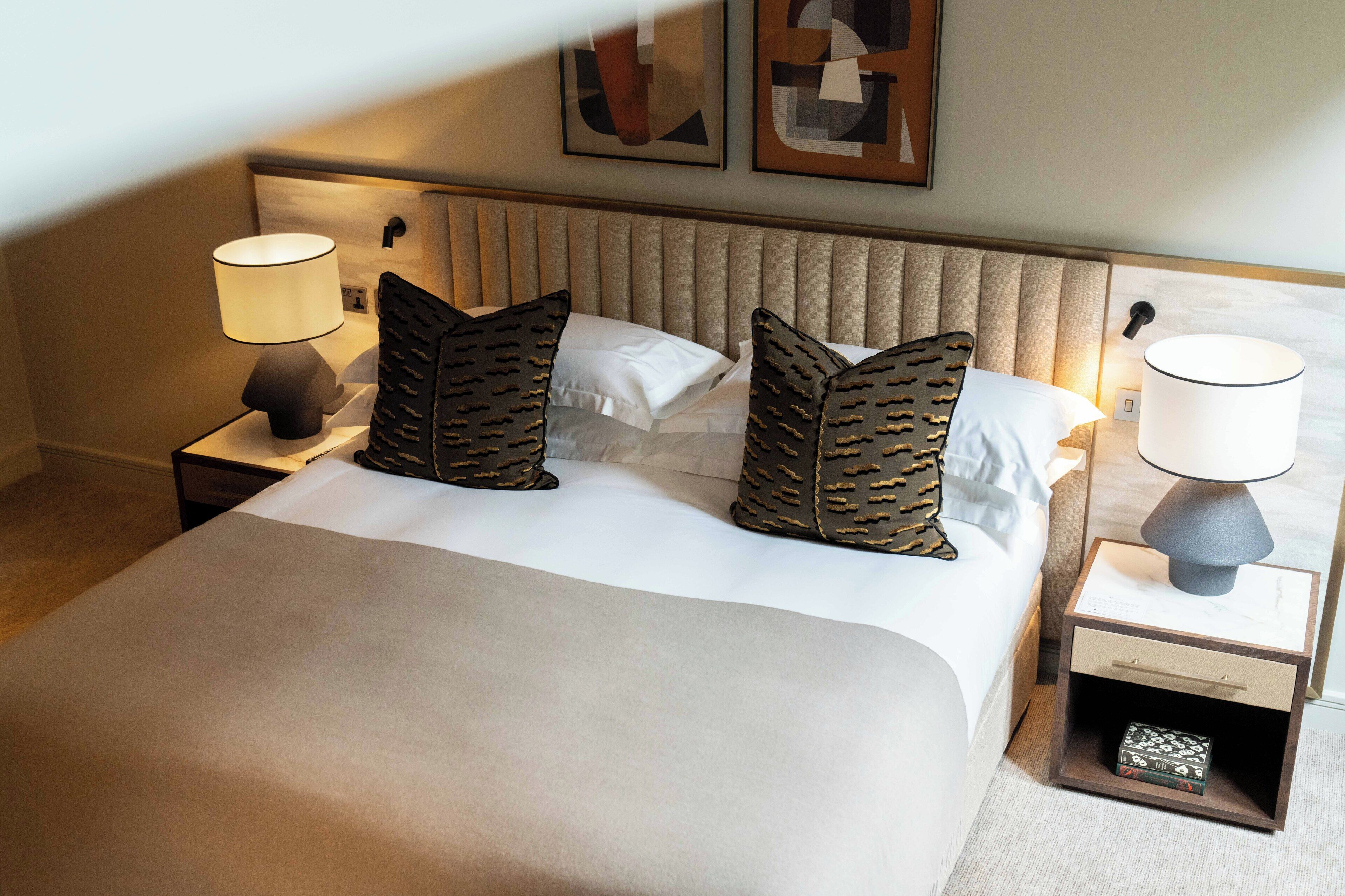 Cama bem arrumada com cabeceira bege, dois travesseiros decorativos e mesa de cabeceira com abajur.