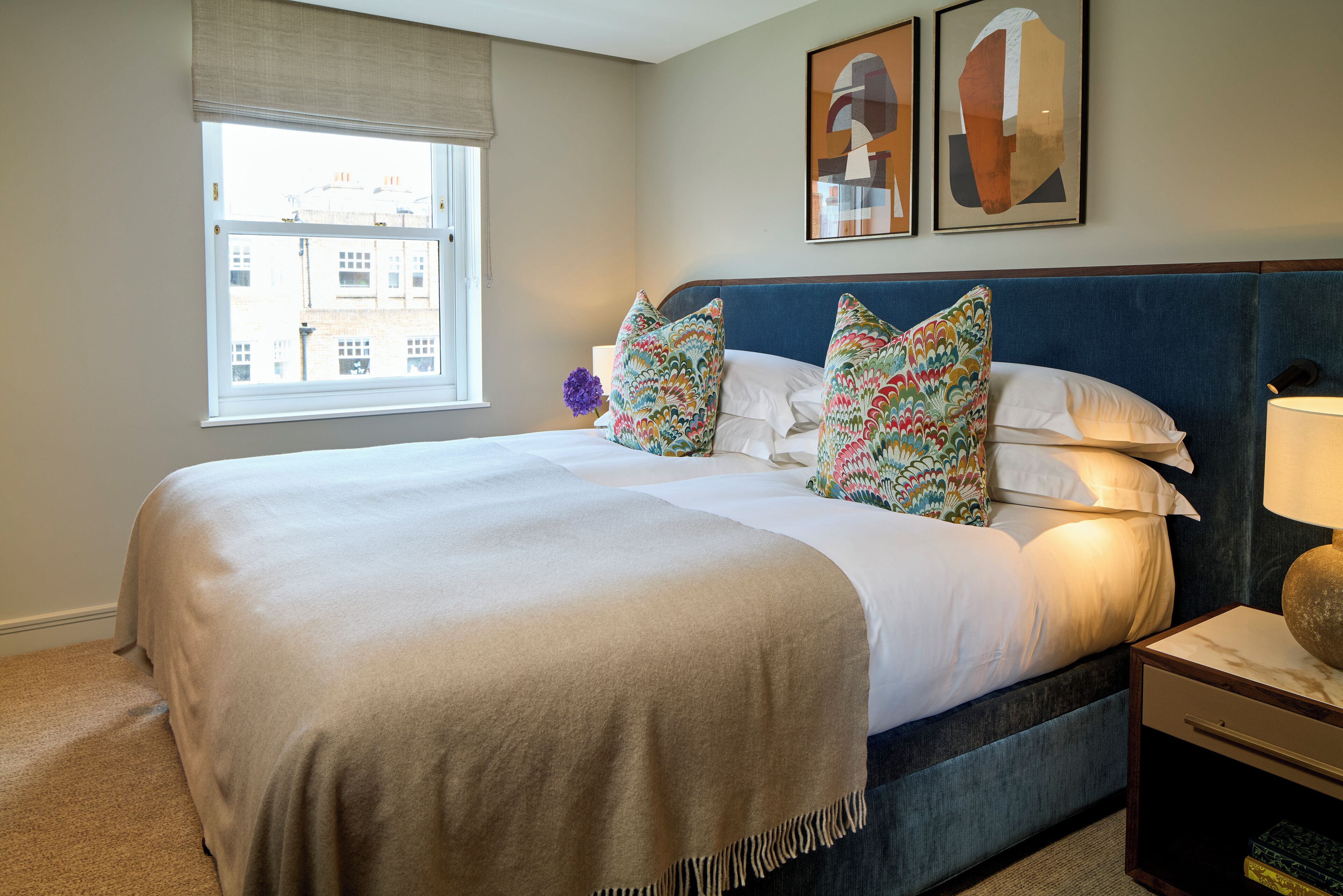 A neatly made bed with a beige blanket and patterned pillows, a bedside table with a lamp, and a window with a view of buildings.
