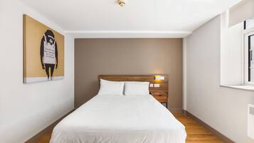 Una cama bien hecha con sábanas blancas en un dormitorio moderno con cabecera de madera, una lámpara de pared y una ventana a la derecha.