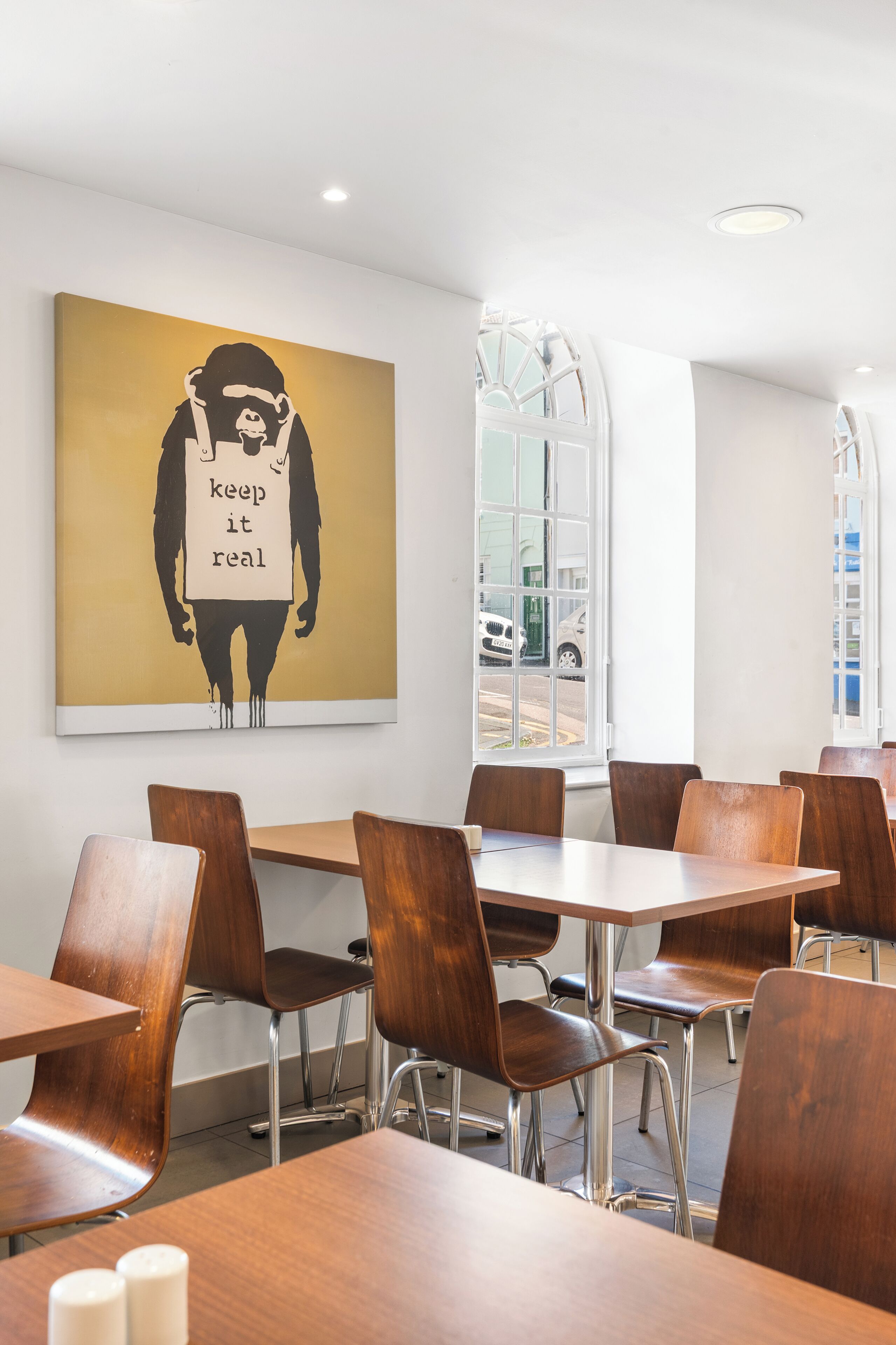 A modern dining area with wooden tables and chairs, a large wall-mounted artwork, and a window with a view of the street.