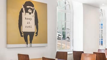 A modern dining area with wooden tables and chairs, a large wall-mounted artwork, and a window with a view of the street.