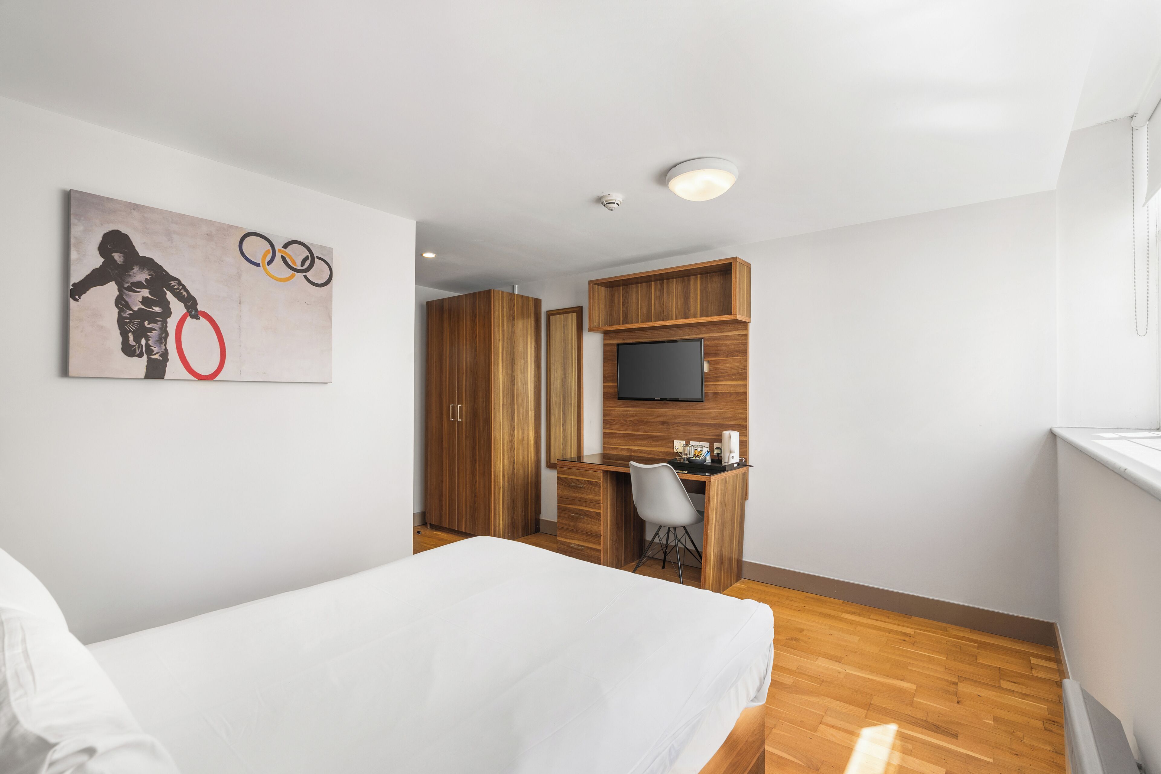 A hotel room with a bed, a desk with a chair, a television, and a painting on the wall.