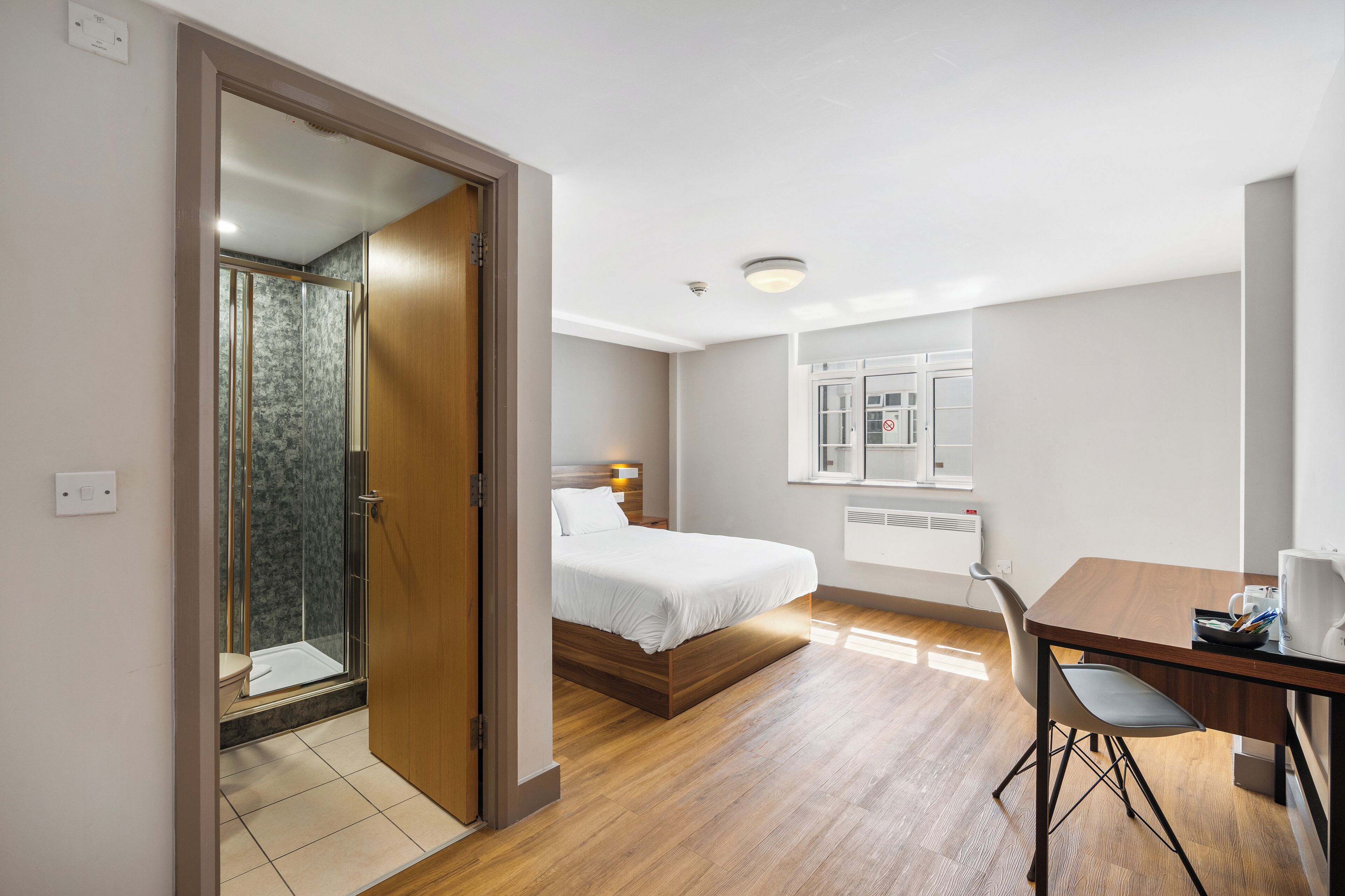 A hotel room with a bed, a desk, a chair, a shower, and a window.