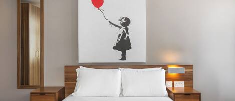 Habitación de hotel con cama, mesita de noche, lámpara de noche y una obra de arte en la pared que muestra a una niña sosteniendo un globo con forma de corazón.