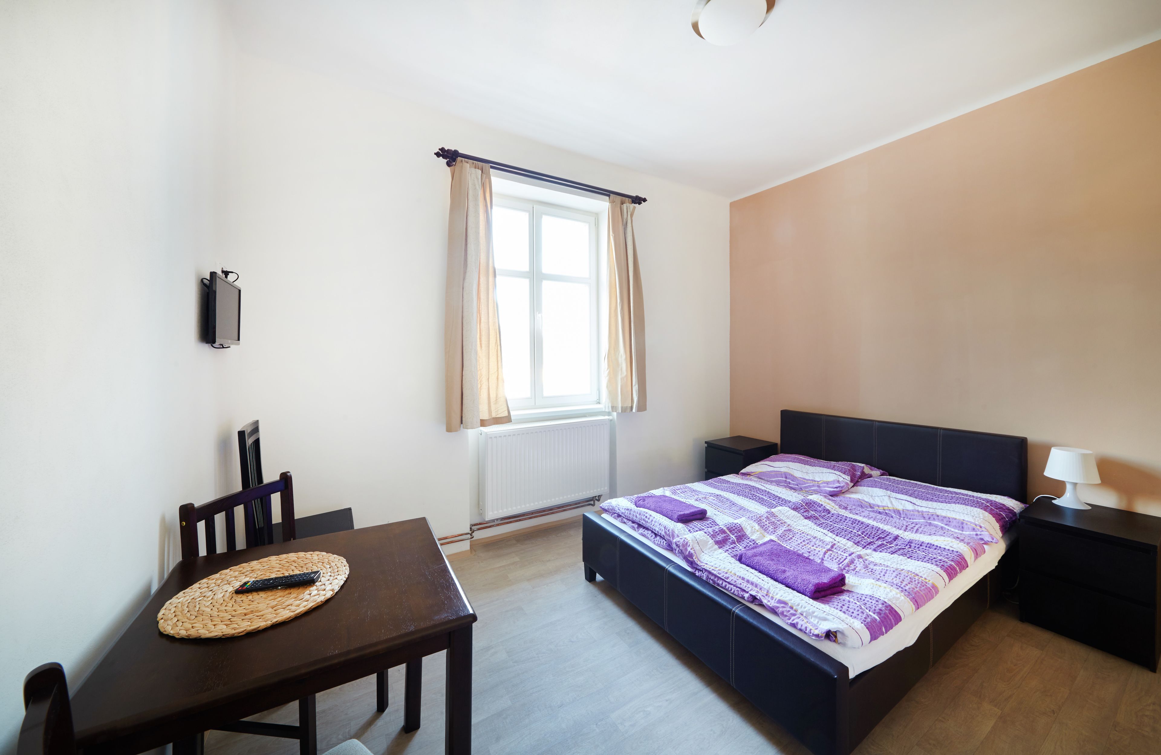 Un dormitorio con una cama, una mesita de noche, una silla, una mesa con un control remoto y una ventana con cortinas.