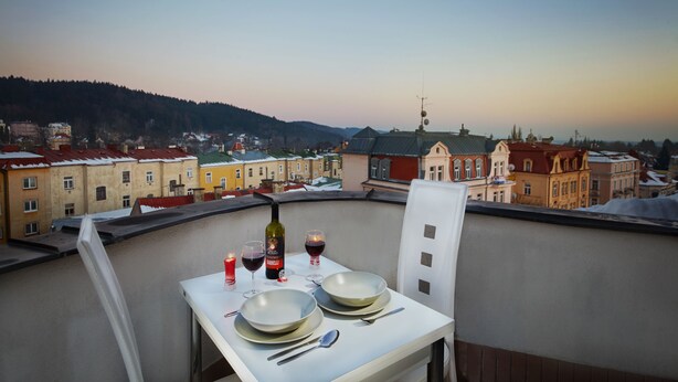 A balcony table set with plates, glasses, and a bottle, overlooking a cityscape with buildings and hills. - Hostel Foster (Marianske Lazne)