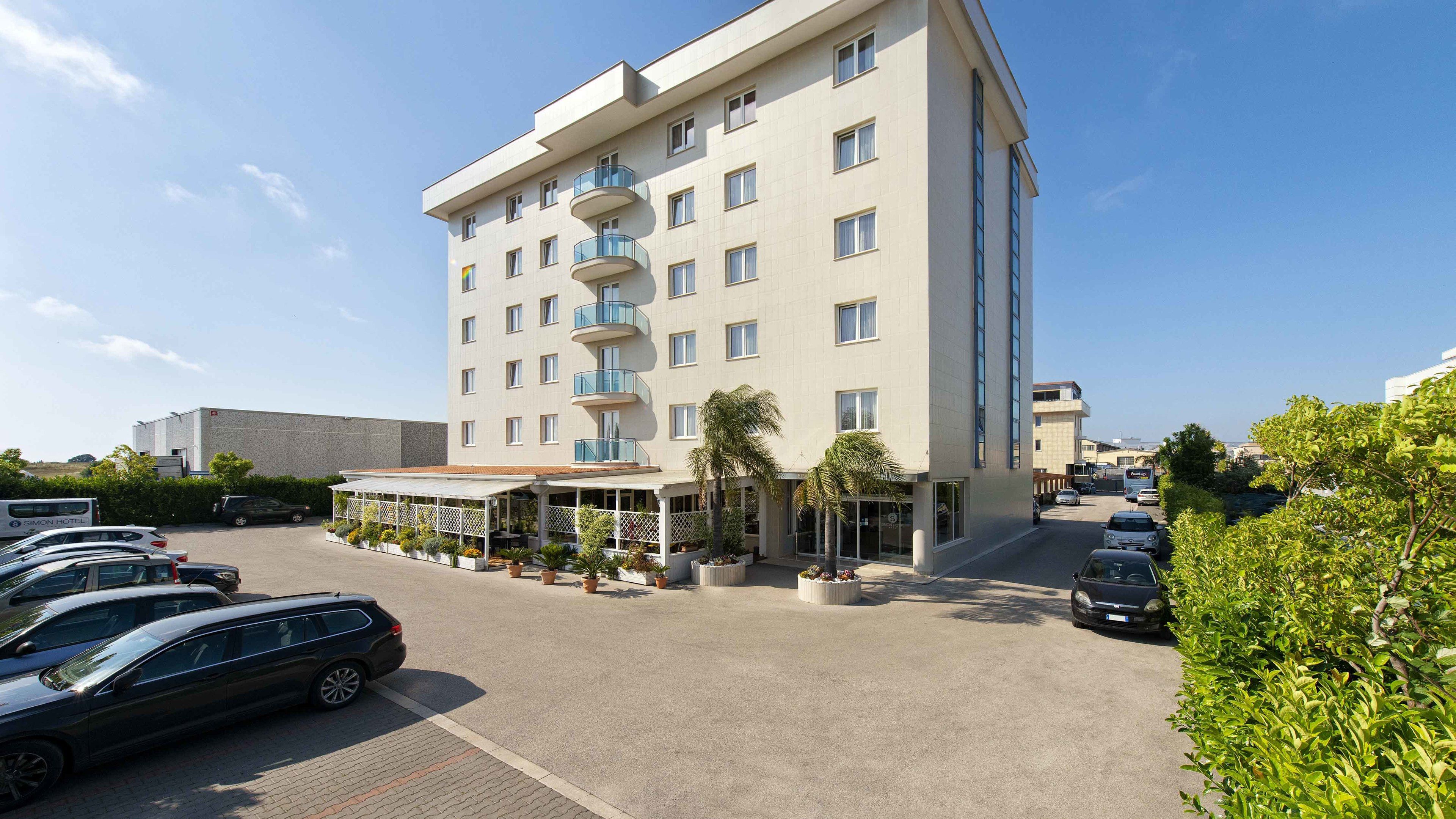 A multi-story hotel with a covered outdoor seating area, a parking lot with several cars, and a clear blue sky.