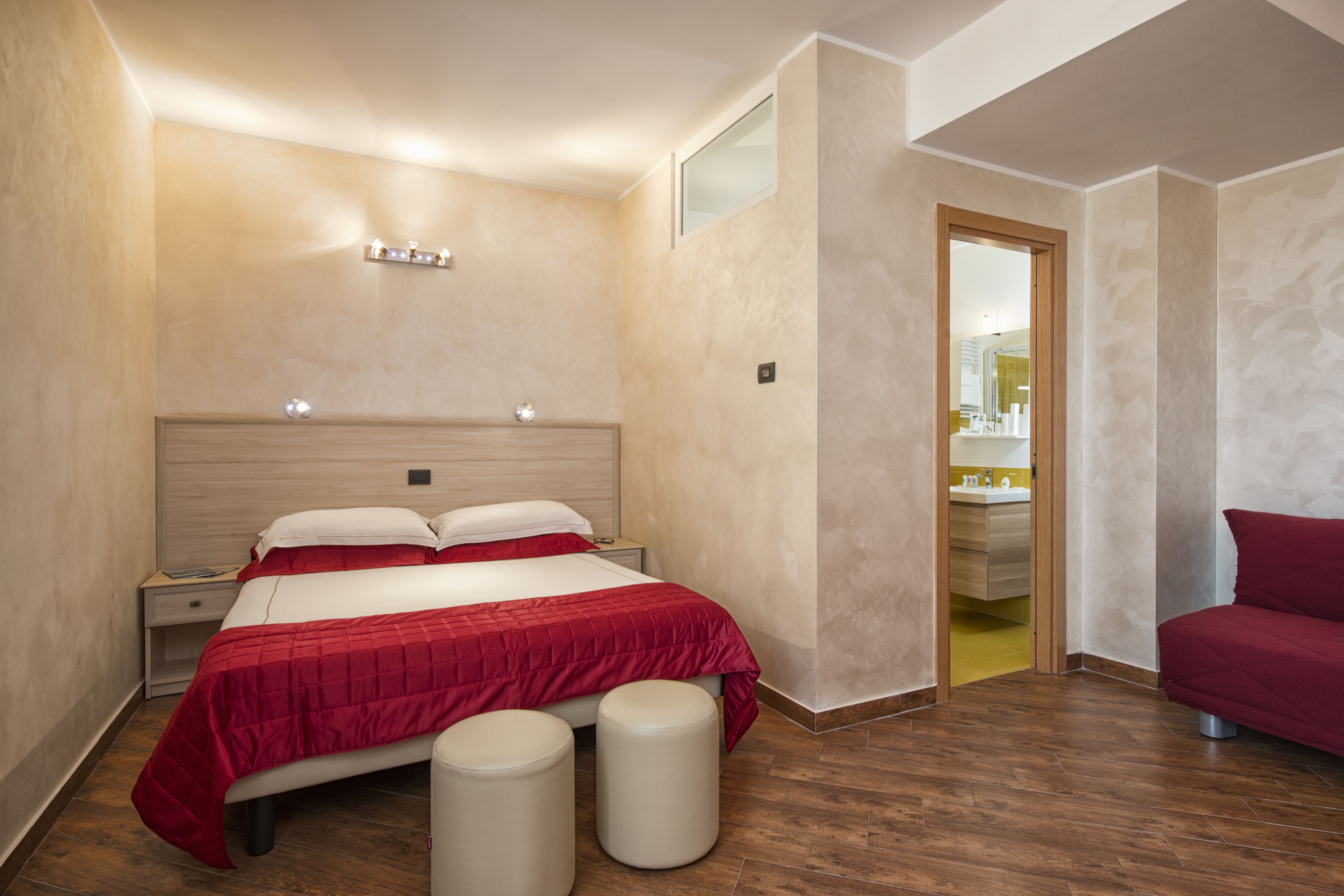 A bedroom with a large bed, two bedside tables, a nightstand, a red bedspread, two white cylindrical stools, and a wooden floor.