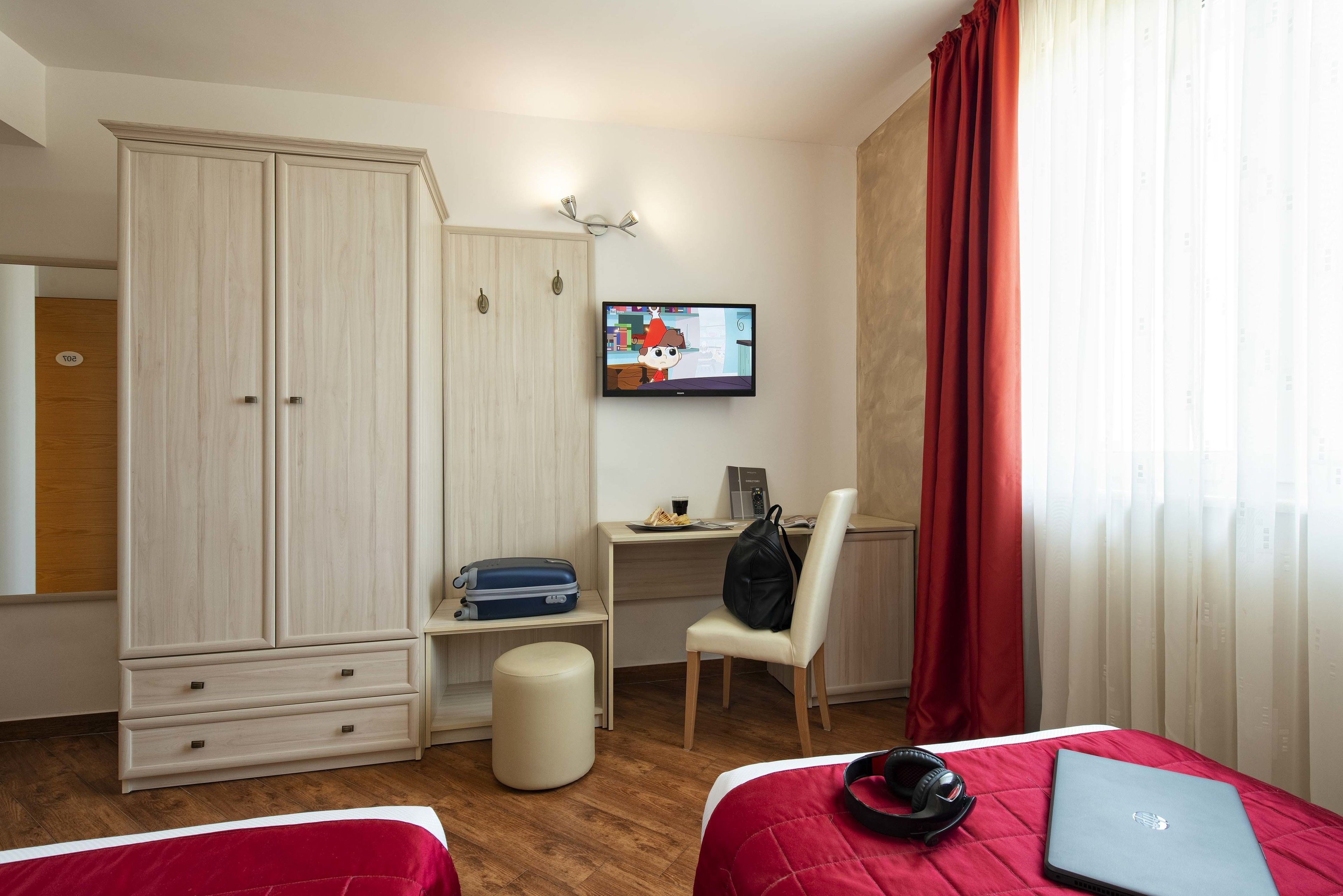 Ein Hotelzimmer mit Bett, Schreibtisch, Laptop, Fernseher, Kleiderschrank und Stuhl.