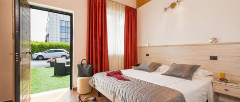 A bedroom with a bed, bedside tables, a nightstand, a window with curtains, a door, and a view of a car and building outside.