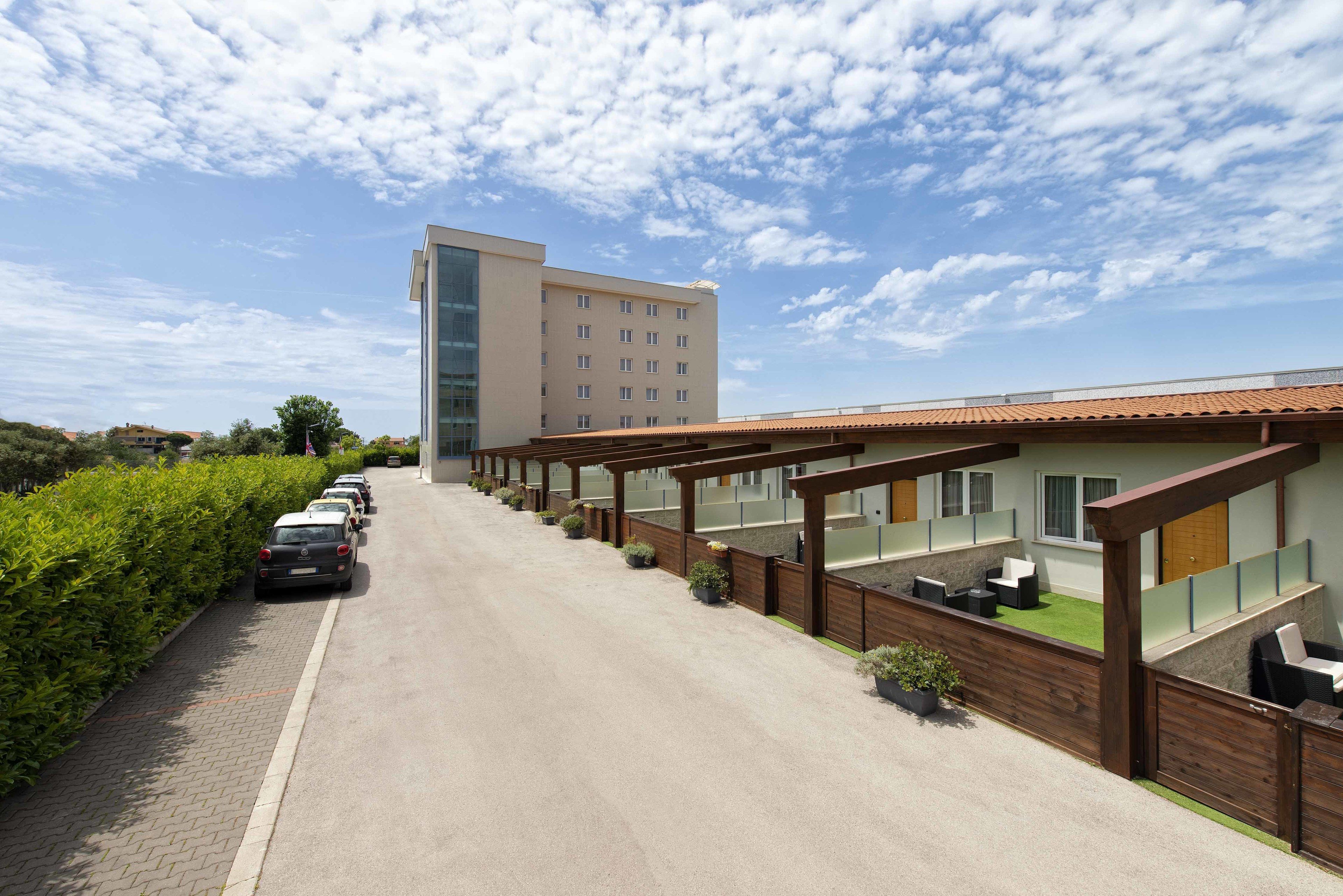 Ein Hotel mit einer Reihe geparkter Autos, einer überdachten Außenbereichssitzgruppe und einem modernen Gebäude im Hintergrund.