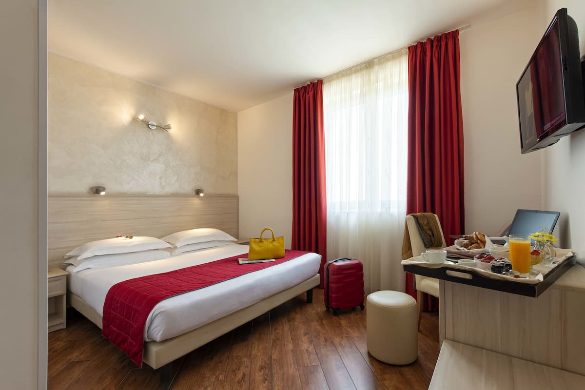 A hotel room with a bed, bedside table, a desk with breakfast, a suitcase, and a TV.