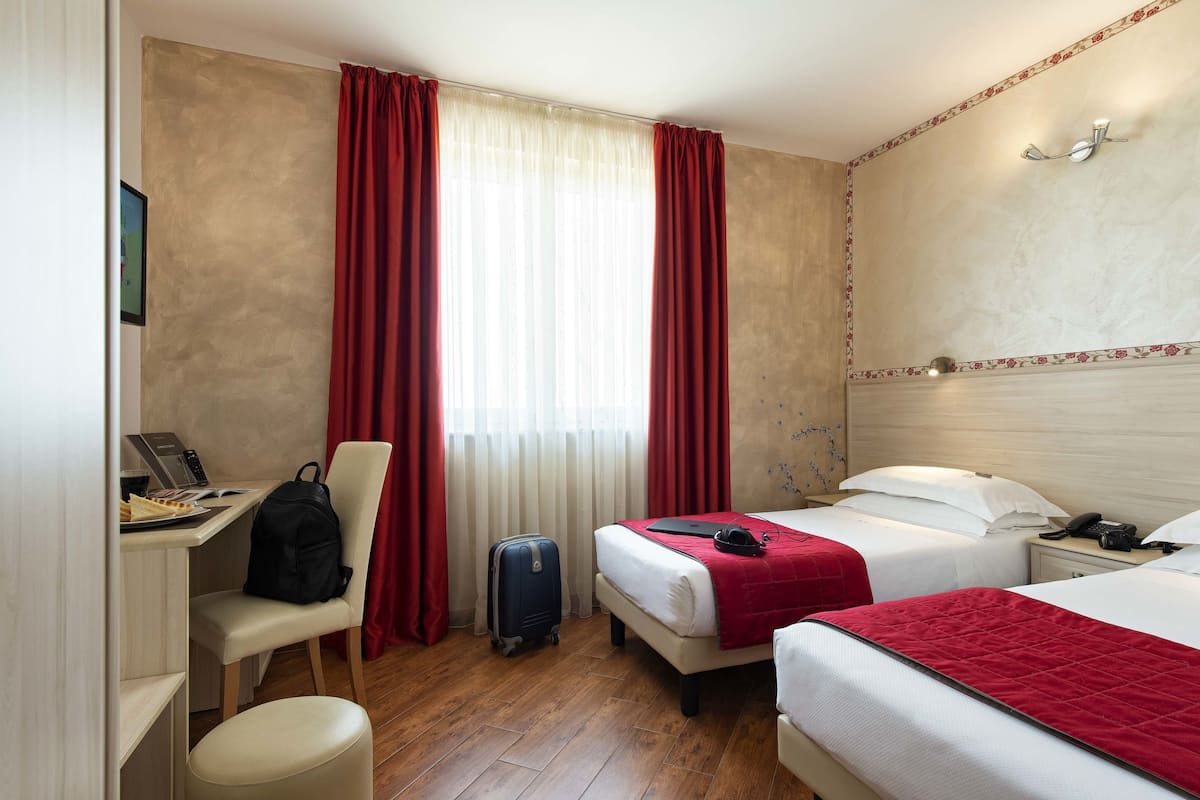 A hotel room with two beds, a desk with a laptop, a chair, a suitcase, and a red curtain.