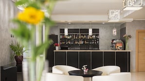 A modern bar area with a counter displaying various bottles, a coffee machine, and a menu board. There are two white chairs in the foreground and a potted plant to the left. - Simon Hotel (Pomezia)