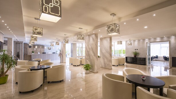A modern hotel lobby with a spacious layout, featuring a reception desk, seating areas, and a visible kitchen in the background. - Simon Hotel (Pomezia)
