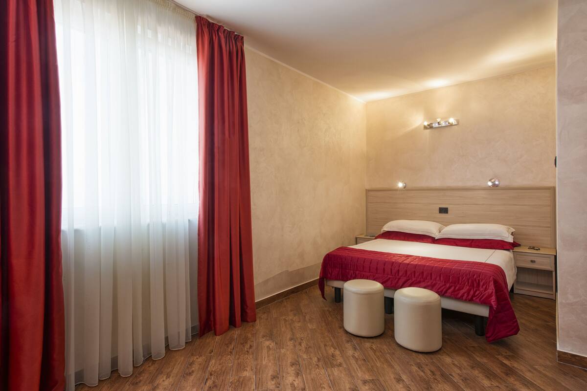 A hotel room with a wooden floor, a bed with red bedding, a bedside table, and a nightstand.