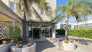 A hotel entrance with a covered walkway, palm trees, and flower planters. - Simon Hotel (Pomezia)