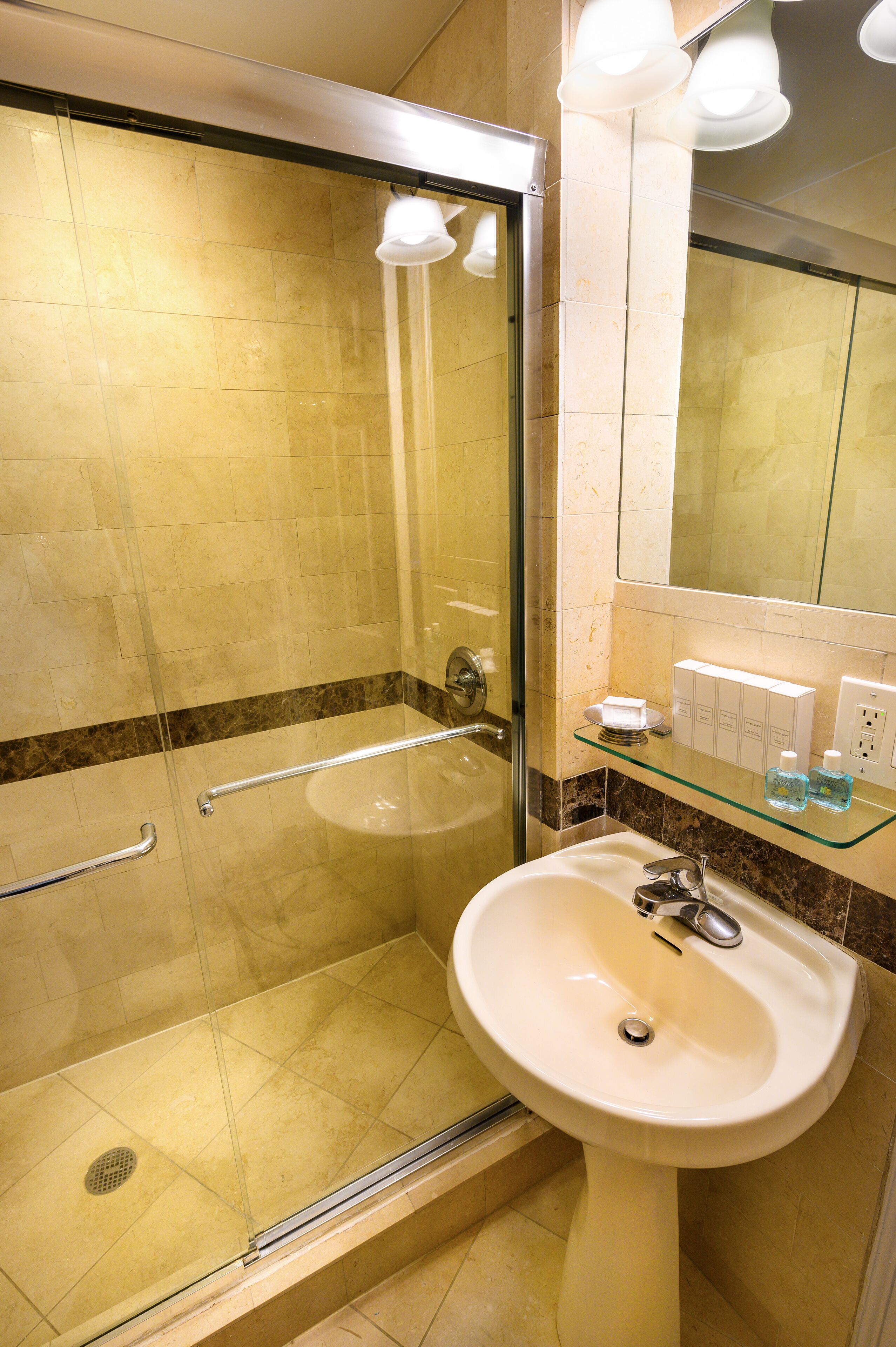 A bathroom with a glass-enclosed shower, a white sink, and a glass shelf with toiletries.