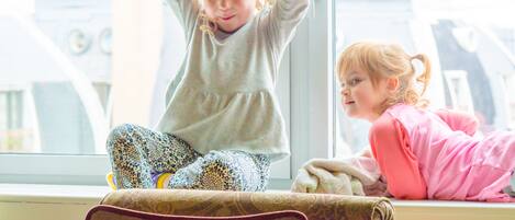 Dos niños jugando debajo de una cama en una habitación con un ventanal grande.