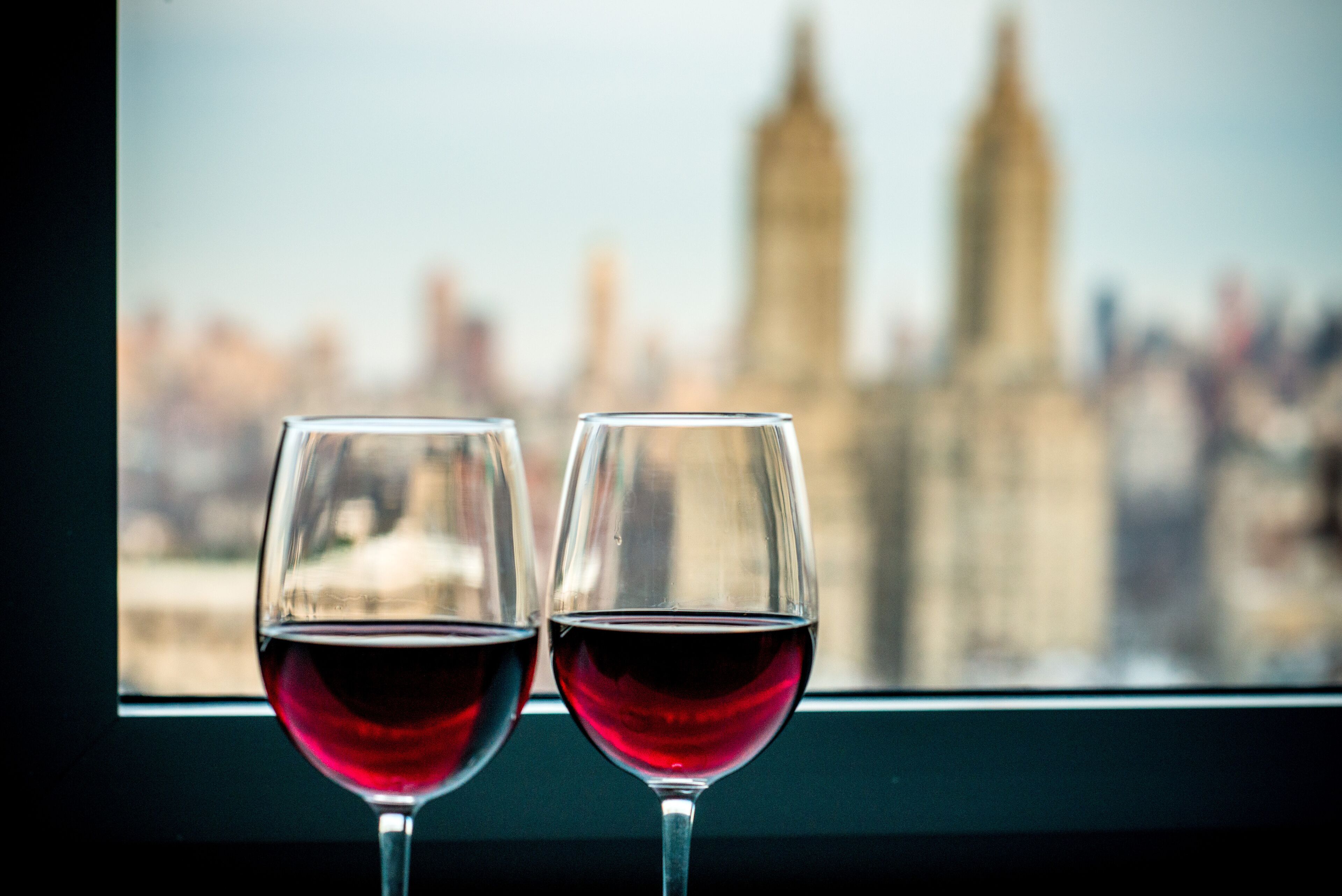 Two wine glasses with red wine on a windowsill, with a blurred cityscape in the background.