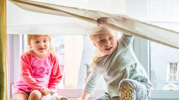 Deux enfants jouent sous une structure ressemblant à une tente, dans une pièce dotée d’une grande fenêtre.
