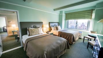 A hotel room with two beds, a desk, a chair, a window with a view of the city, and a bathroom visible through an open door.
