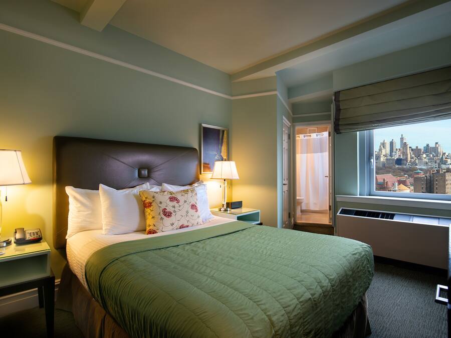 A hotel room with a bed, two bedside tables with lamps, a city view through the window, and a door leading to another room.