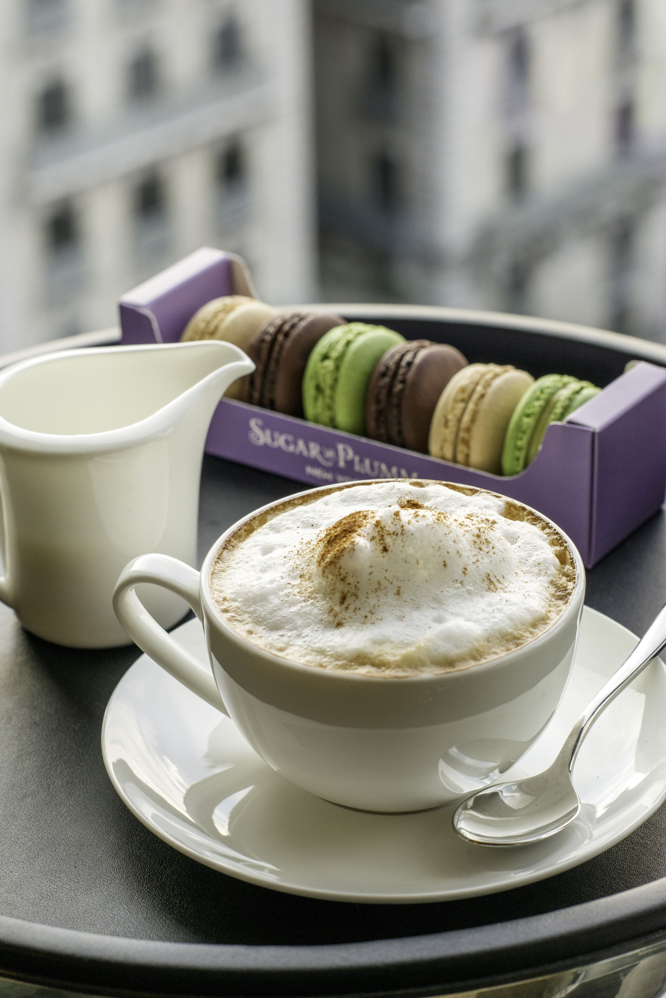 A cup of coffee with foam and a sprinkle of cinnamon, a white pitcher, and a box of colorful macarons on a table.