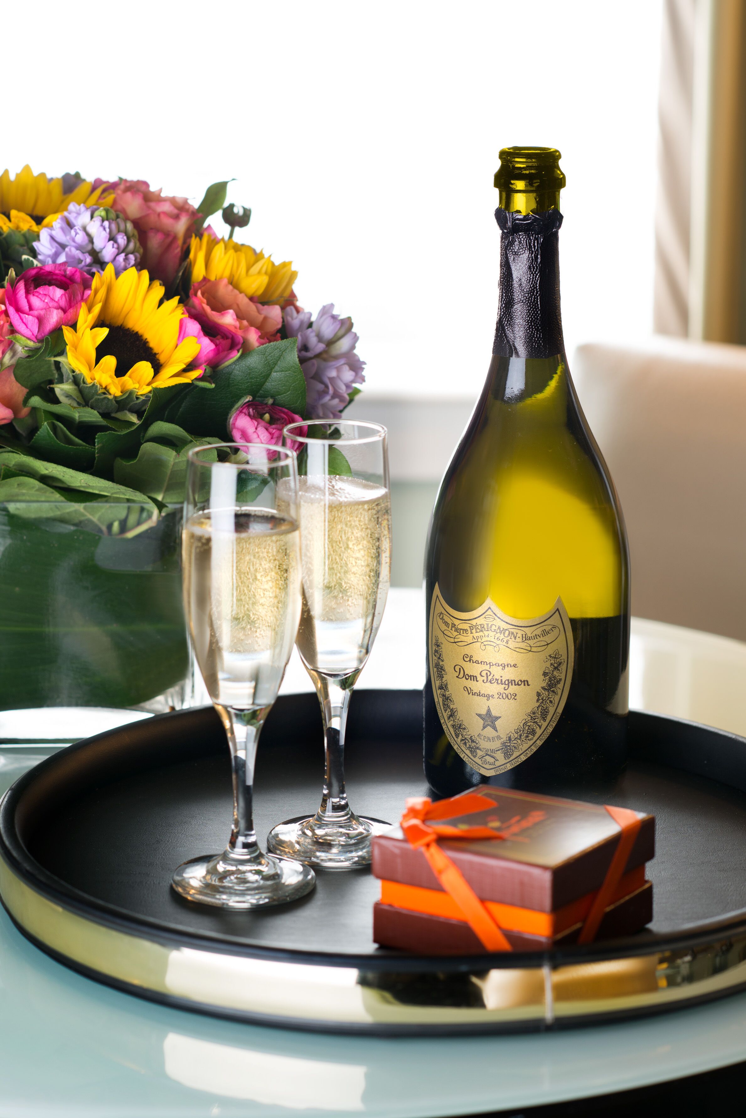 Une bouteille de champagne Dom Pérignon, deux coupes de champagne, un bouquet de fleurs et deux petites boîtes à cadeaux sur un plateau.