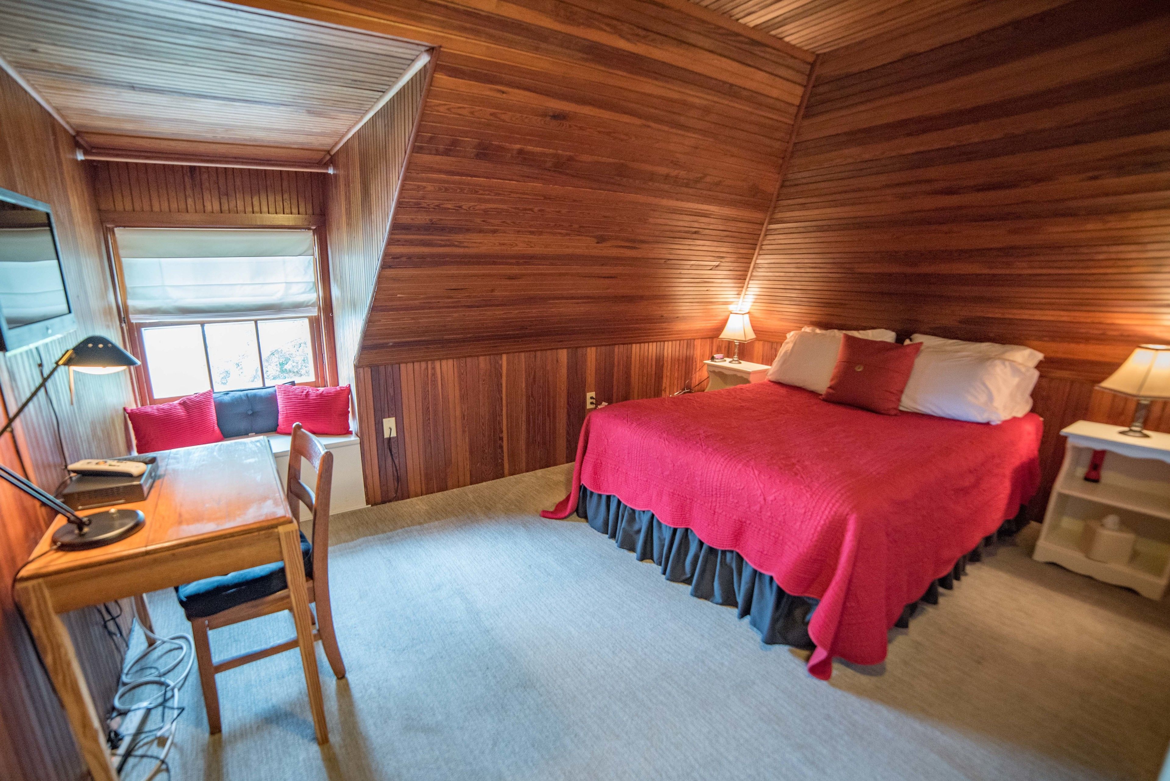 Une chambre confortable avec un plafond en bois, un lit recouvert d’une courtepointe rouge, un bureau avec une lampe et une fenêtre équipée de stores.