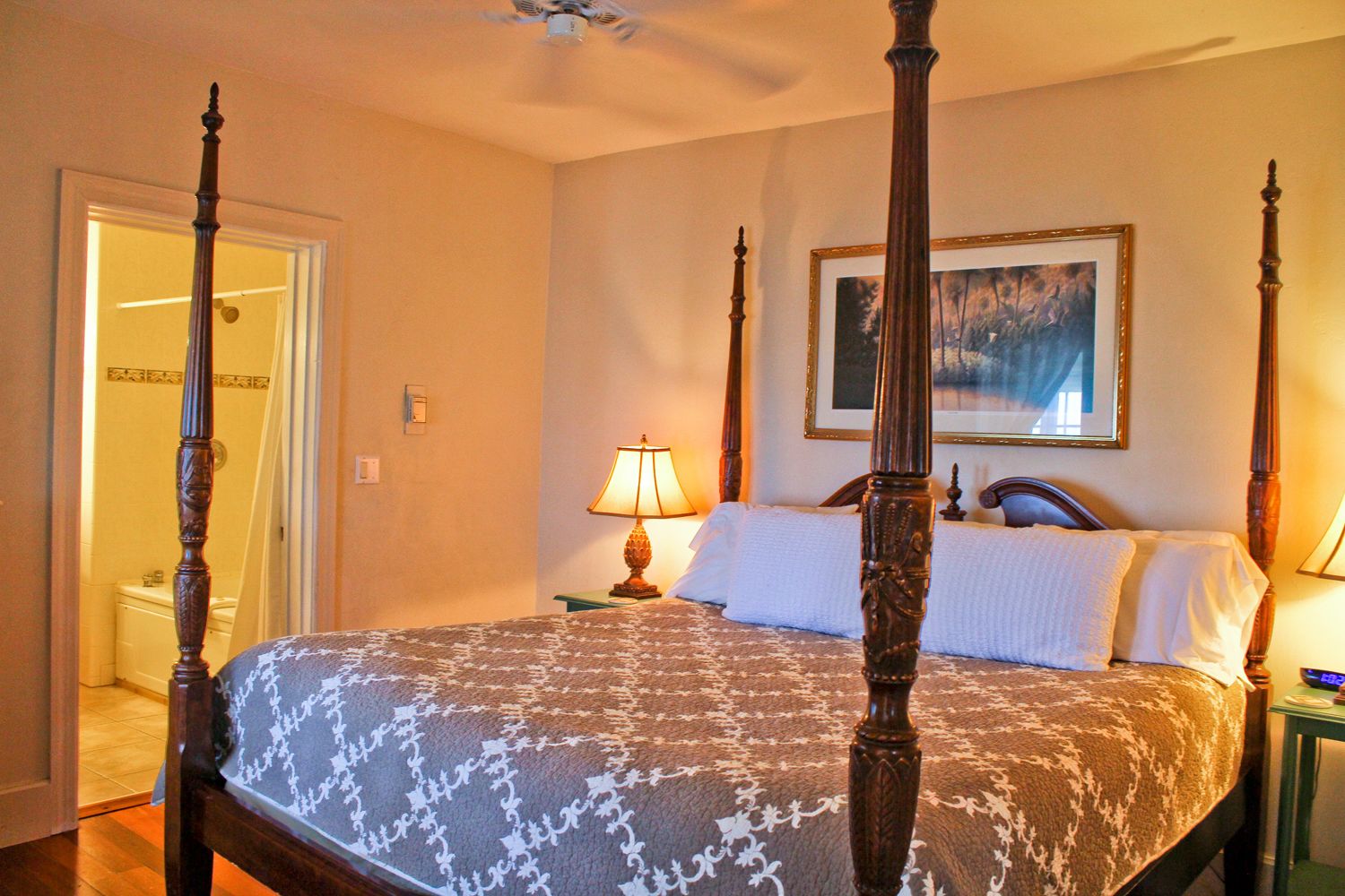 A four-poster bed with a patterned bedspread, a bedside lamp, and a framed picture on the wall.