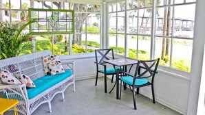 Een zonnedoorspoelde kamer met een witte rieten bank, kussens in teal en een kleine tafel met stoelen.