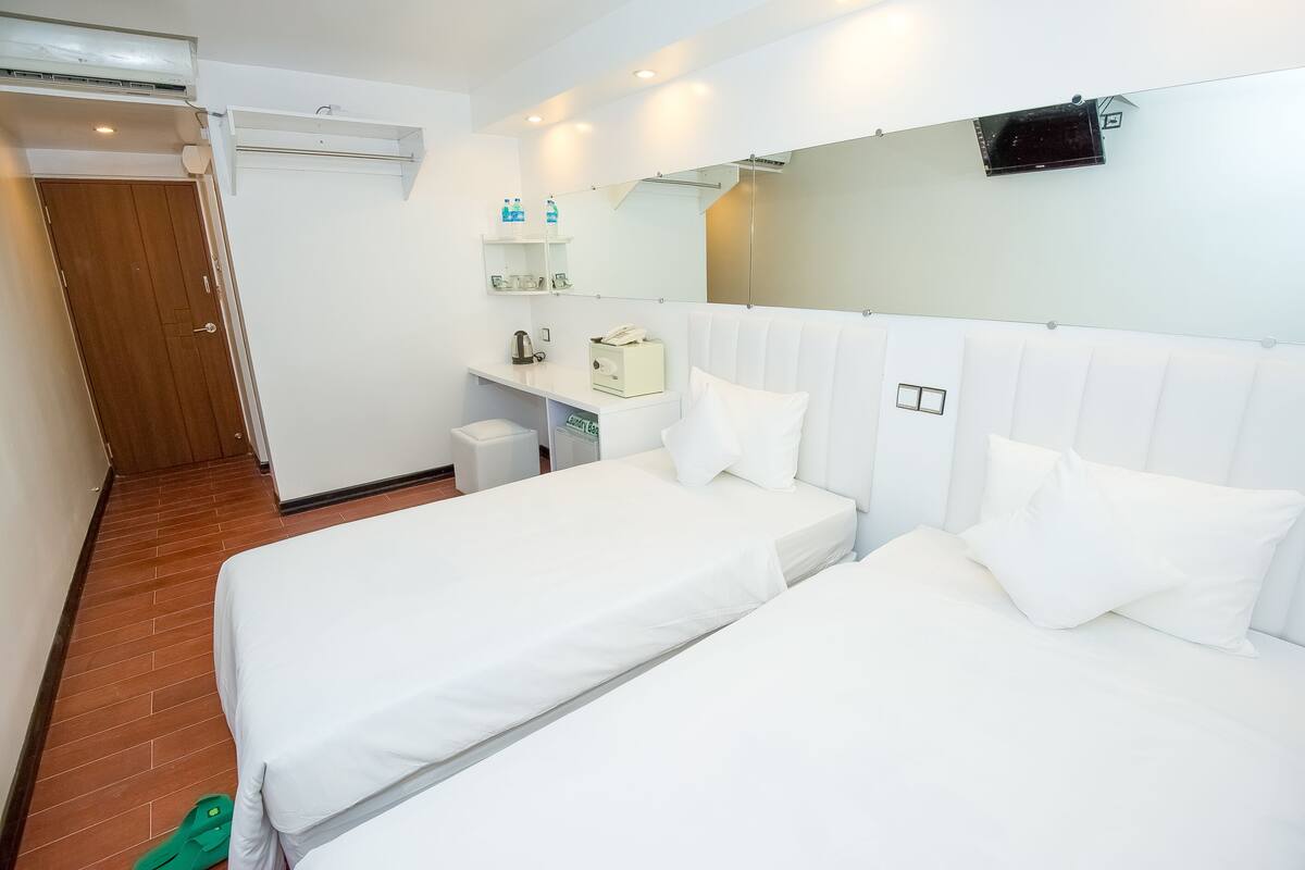 A hotel room with two single beds, a small desk with a chair, a mirror, and a shelf with toiletries.
