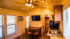 L’intérieur d’une cabane en bois comprend une table à manger, des chaises, un coin cuisine et un ventilateur de plafond.