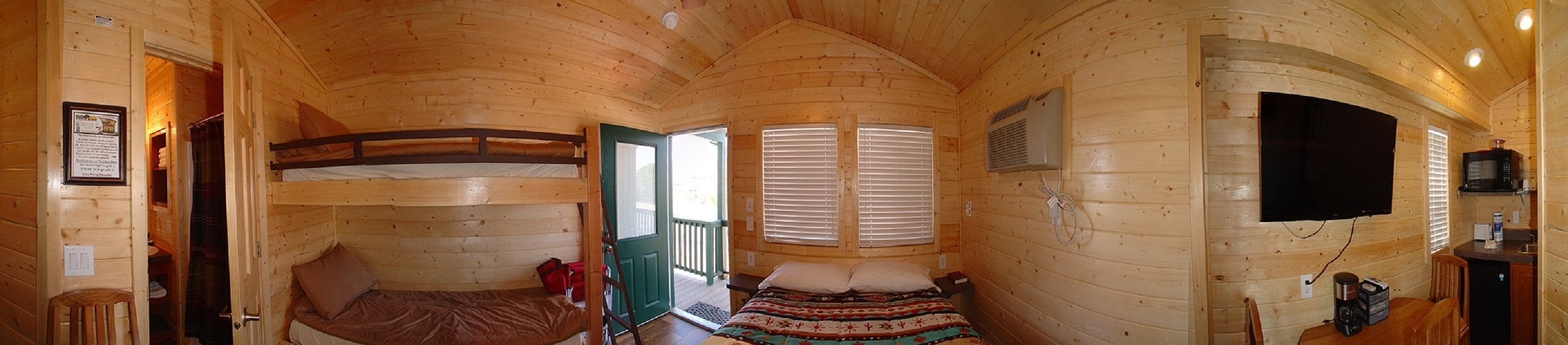 A cabin with two bunk beds, a small kitchenette, a TV, and a window with blinds.